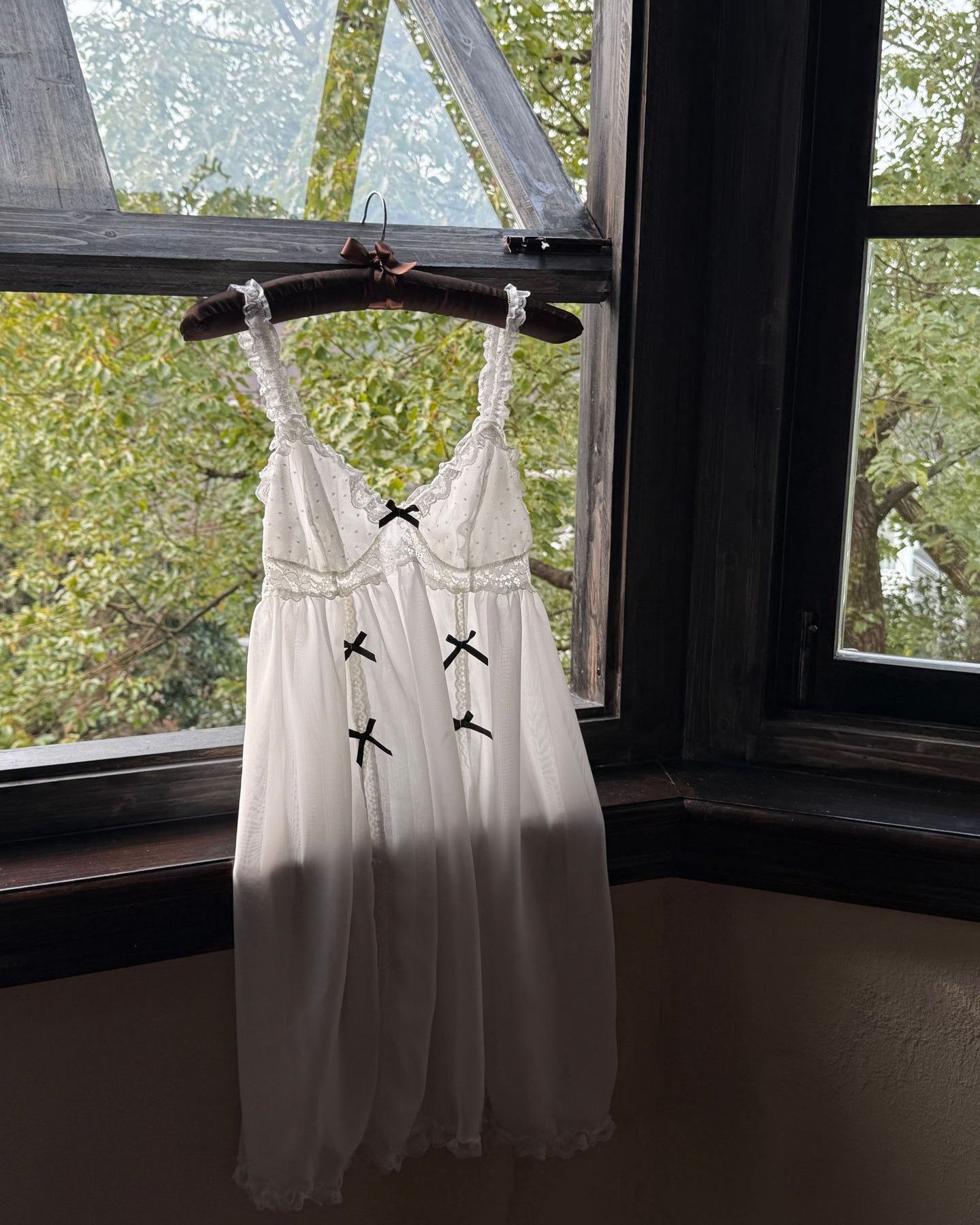White lace dress with black accents on a hanger in front of a window with greenery outside.