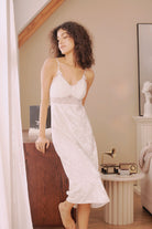 Side profile of model in the white velvet nightdress, standing by a window. Natural light enhances the texture and soft glow of the dress.