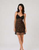 A woman modeling a short black and brown leopard-print lingerie chemise with a black lace bust and thin straps, standing against a light gray studio background.