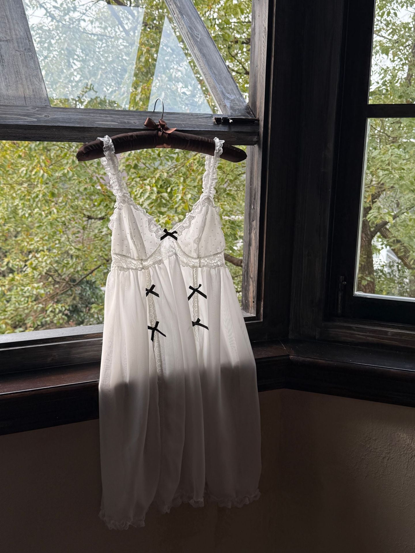 White lace dress with black accents on a hanger in front of a window with greenery outside.